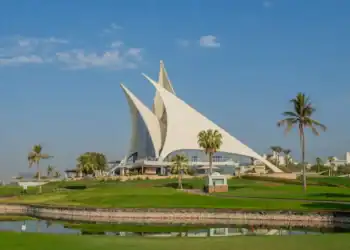 The iconic sail-shaped clubhouse of Dubai Creek Golf & Yacht Club on a sunny day, with lush green fairways, a water hazard in the foreground, and palm trees under a blue sky.