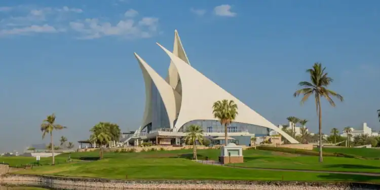 The iconic sail-shaped clubhouse of Dubai Creek Golf & Yacht Club on a sunny day, with lush green fairways, a water hazard in the foreground, and palm trees under a blue sky.