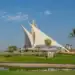 The iconic sail-shaped clubhouse of Dubai Creek Golf & Yacht Club on a sunny day, with lush green fairways, a water hazard in the foreground, and palm trees under a blue sky.