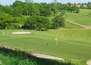 This image shows a smoothly contoured putting green in the foreground, bordered by a bunker, with gently rolling fairways and another bunker visible further back.