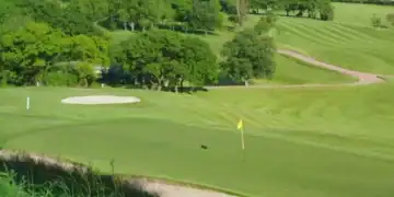 This image shows a smoothly contoured putting green in the foreground, bordered by a bunker, with gently rolling fairways and another bunker visible further back.
