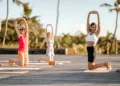 A group of people doing a kneeling yoga stretch on an outdoor wooden deck surrounded by palm trees and greenery under a blue sky at Heritage Resorts & Golf in Mauritius.