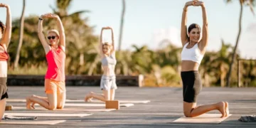 A group of people doing a kneeling yoga stretch on an outdoor wooden deck surrounded by palm trees and greenery under a blue sky at Heritage Resorts & Golf in Mauritius.