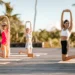 A group of people doing a kneeling yoga stretch on an outdoor wooden deck surrounded by palm trees and greenery under a blue sky at Heritage Resorts & Golf in Mauritius.